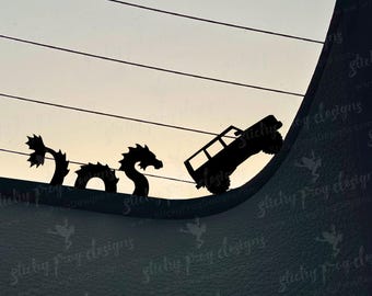 May include: Black silhouette decal featuring a dragon and a 4x4 vehicle climbing a hill. The dragon has a long, serpentine body and a detailed head. The vehicle is a classic off-road style. The background is a gradient of light and dark.