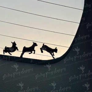 May include: Silhouette of three goats in mid-jump against a gradient sky. The goats are black, with visible details. The background features horizontal lines and a curved dark border, creating a dynamic and playful scene.