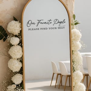 Op de afbeelding: Een goudomrandde spiegel met de tekst "Our Favorite People, Please Find Your Seat" in zwart script. De spiegel is versierd met witte bloemen en groen, wat duidt op een bruiloft of evenement.