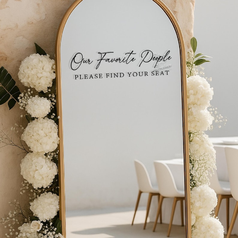 Op de afbeelding: Een goudomrandde spiegel met de tekst "Our Favorite People, Please Find Your Seat" in zwart script. De spiegel is versierd met witte bloemen en groen, wat duidt op een bruiloft of evenement.