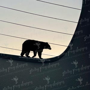 May include: A black silhouette of a bear walking across a curved surface, set against a gradient sky. The image includes the text "ONLY PURCHASE on ETSY @ STICKYFROGDESIGNS.COM". The design is likely a decal or sticker.