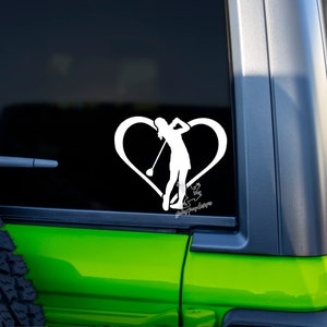 May include: White vinyl decal of a golfer swinging a club, set within a heart shape. The silhouette of the golfer is detailed, with the club raised in a swing. The decal is applied to a vehicle window.