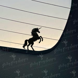 May include: Black silhouette of a rearing unicorn with a horn, set against a gradient sky. The unicorn is a sticker or decal, likely for a car window. The background has horizontal lines and the text "Sticky Frog Designs".