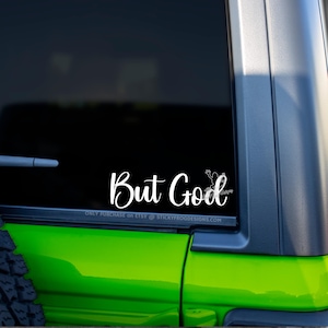 May include: White vinyl decal with the words "But God" in a script font, accompanied by a small frog illustration. The decal is affixed to the rear window of a green vehicle.