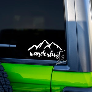 May include: White vinyl decal with a mountain range silhouette and the word "wanderlust" in a cursive font. The decal is on the back window of a green car.