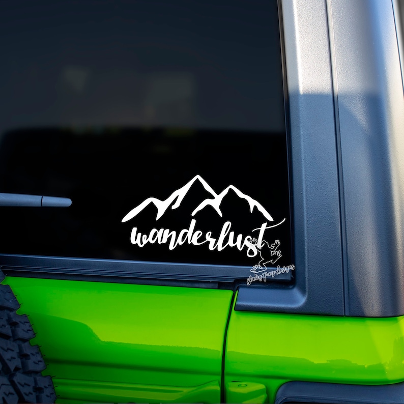 May include: White vinyl decal with a mountain range silhouette and the word "wanderlust" in a cursive font. The decal is on the back window of a green car.