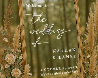 May include: A decorative mirror with a gold frame displays wedding details. The text reads "Welcome to The wedding of NATHAN & LANEY OCTOBER 4, 20XX We're so glad you're here." The mirror is surrounded by floral arrangements.