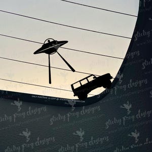 May include: Black vinyl decal featuring a UFO abducting a 4x4 vehicle. The design includes the text "ONLY PURCHASE on ETSY @ STICKYFROGDESIGNS.COM". The background shows a gradient sky.