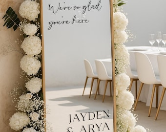 May include: A gold-framed, arched mirror with the text "We're so glad you're here" and names "JAYDEN & ARYA" with a date. The mirror is decorated with white flowers and greenery, suitable for a wedding or event.