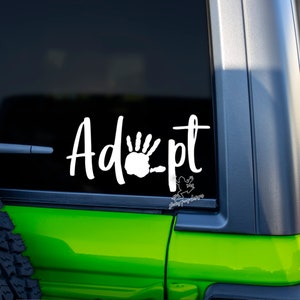 May include: White vinyl decal with the word "Adopt" in a cursive font. A white handprint is in the middle of the word. The decal is on a green car window.