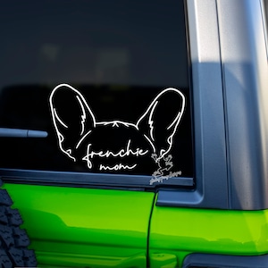 Peut inclure: Autocollant en vinyle blanc représentant la tête d'un bouledogue français avec le texte "Frenchie Mom" sur la vitre d'une voiture verte.