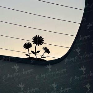 May include: Silhouette of three daisy-like flowers with stems and leaves against a light background. The flowers are black, and the background has horizontal lines. The image is a decal or sticker.