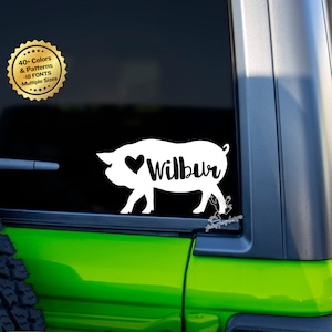 Peut inclure: Autocollant en vinyle blanc représentant un cochon avec un cœur et le nom Wilbur. L'autocollant est sur la vitre d'une voiture verte.