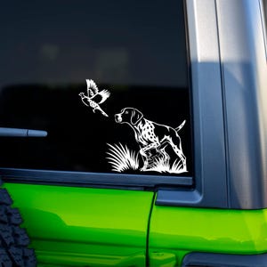 May include: A white decal featuring a hunting dog, a bird in flight, and tall grass. The dog is in a hunting stance, with a bird flying above it. The decal is on a black background, with a green vehicle visible.