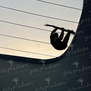 May include: A black silhouette of a sloth hanging from a branch. The image is set against a gradient background of white and gray, with horizontal lines. The design is likely a decal or sticker.