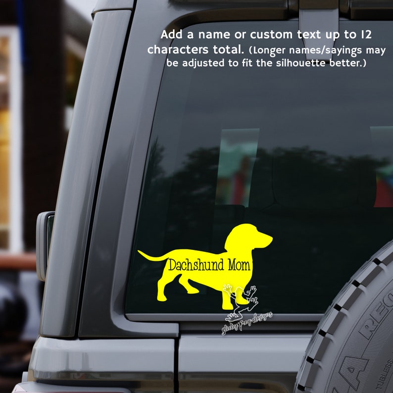 以下が含まれることがあります： 黒い車の窓に貼られた、ダックスフントのシルエットと「Dachshund Mom」の文字が書かれた黄色のビニールステッカー。背景にはタイヤが見える。