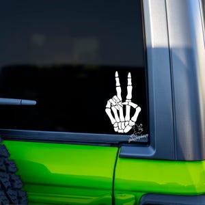 May include: White skeleton hand making the peace sign. The hand is on a green car window with a black frame.