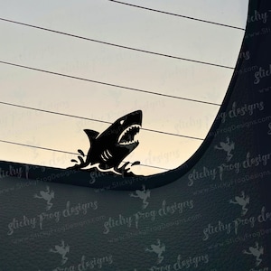May include: A black shark decal with an open mouth and visible teeth, emerging from stylized water waves. The decal is designed to be applied to a car window or similar surface. The background shows a gradient of colors.