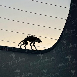 May include: A black silhouette of a snarling, spiky-backed creature, possibly a wolf or dog, is depicted against a gradient sky. The creature is positioned on a curved surface, suggesting a sticker or decal. The background has horizontal lines.