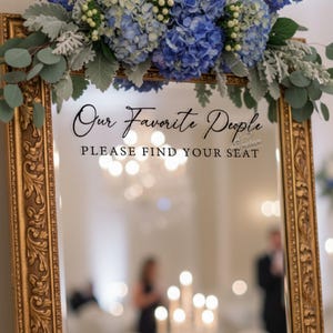 May include: A decorative mirror with a gold ornate frame, featuring the text "Our Favorite People, Please Find Your Seat." The mirror is adorned with blue and white flowers and greenery, suitable for a wedding or special event.