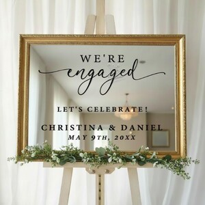 May include: A gold framed mirror with the words "WE'RE engaged" in elegant script. Below, it reads "LET'S CELEBRATE!" followed by names and a date. The mirror is decorated with greenery and rests on a wooden easel.