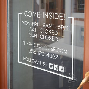 May include: A white rectangular sign with rounded corners on a glass door. The sign says 'Come Inside!' and lists business hours: 'Mon-Fri 9am-5pm, Sat Closed, Sun Closed.' The sign also includes the business name 'ThePhotoHouse.com', phone number '555-123-4567', and social media handles: 'Follow Us: Twitter, Instagram, Facebook.'
