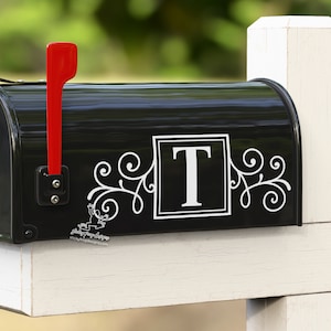 May include: Black mailbox with a white letter "T" in a square frame and decorative swirls on either side. The mailbox has a red flag.