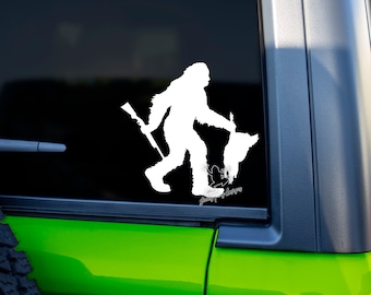 Op de afbeelding: Witte silhouet van een Bigfoot die een geweer en een kalkoen vasthoudt. De Bigfoot loopt naar rechts. De afbeelding is op een groene auto.