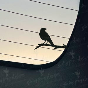 May include: Black silhouette of a crow perched on a branch, set against a gradient sky. The crow is detailed, with visible features. The design is likely a decal or sticker, suitable for car windows or similar applications.