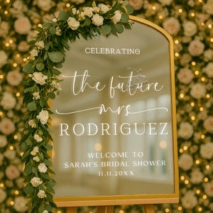 May include: A gold-framed mirror sign with the words "Celebrating the future Mrs. Rodriguez" in elegant white script. The sign welcomes guests to Sarah's bridal shower. The mirror is decorated with white roses and greenery.