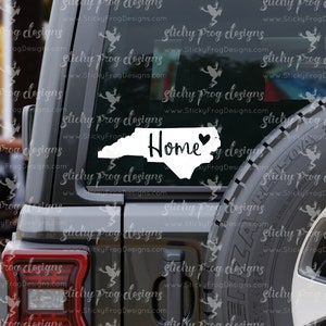 May include: A white vinyl decal of the state of North Carolina with a heart in the center and the word "Home" written in a cursive font. The decal is on the back window of a gray SUV.