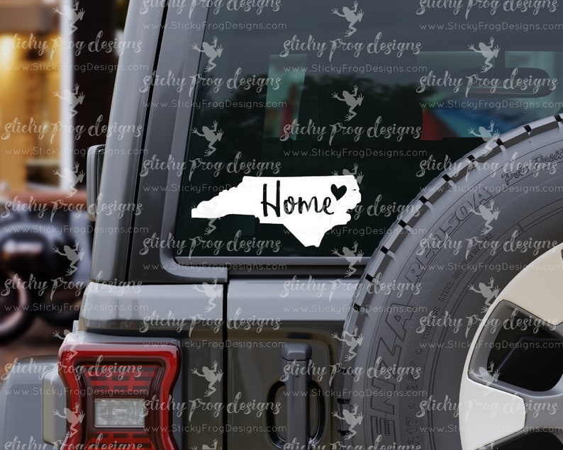 May include: A white vinyl decal of the state of North Carolina with a heart in the center and the word "Home" written in a cursive font. The decal is on the back window of a gray SUV.