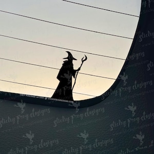 May include: A black silhouette of a wizard holding a staff, set against a light background with horizontal lines. The wizard wears a pointed hat and a long robe. The image includes the text "ONLY PURCHASE on ETSY @ STICKYFROGDESIGNS.COM".