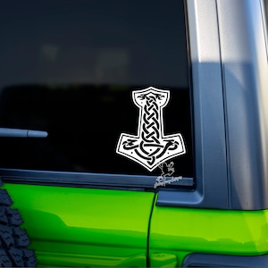 May include: A white vinyl sticker of a Mjolnir, Thor's hammer, with a Celtic knot design. The sticker is on the back window of a green car.
