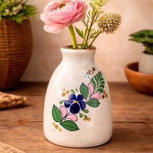 May include: A small, off-white ceramic vase with a rounded body and a narrow neck, featuring a hand-painted floral design in blue, green, and pink. The vase contains a single pink ranunculus, a yellow wildflower sprig, and a dried thistle.