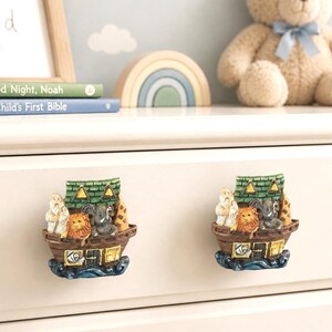 May include: Two drawer knobs shaped like Noah's Ark, featuring a brown boat hull, a green roof, and sculpted figures of a bearded person, a lion, and an elephant. The base of each knob is painted with blue waves, and the sides show yellow windows.
