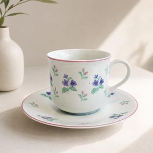 May include: A white porcelain teacup and saucer set featuring a delicate pattern of small purple violet flowers and pink flower buds with green leaves. The rim of both the cup and the saucer is accented with a thin, soft pink painted line.