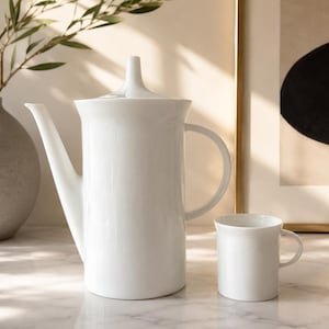 May include: A tall, white ceramic teapot with a slender spout, a matching lid with a pointed finial, and a curved handle, positioned next to a small, matching white ceramic mug with a simple handle, both featuring a smooth, glossy finish.