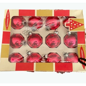 May include: A box of 12 red glass Christmas ornaments. The box is red and gold with the words 'Glass Products' and 'American Made' printed on it. The ornaments are in a clear plastic tray and are arranged in a grid pattern. Each ornament has a silver hanger.