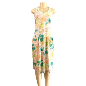 May include: A white sleeveless dress with a floral print in shades of pink, green, and yellow. The dress has a gathered waist and two large pockets on the front.
