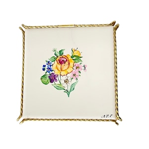 Puede incluir: Una caja de cerámica blanca cuadrada con un marco de metal dorado. La caja presenta un diseño floral pintado a mano con una rosa amarilla, flores moradas, rosas y azules. La caja está firmada "A.M."