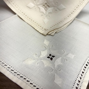 Puede incluir: Dos servilletas cuadradas de lino blanquecino con diseños florales bordados. Una servilleta está doblada, revelando un borde decorativo. La otra servilleta es blanca con un diseño similar. Las servilletas tienen una estética delicada y detallada.