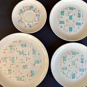 Puede incluir: Un juego de cuatro platos de cerámica vintage y un platillo. Los platos y el platillo son de color blanquecino con una textura moteada y un patrón geométrico de cuadrados turquesa, líneas granates y signos más blancos.