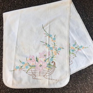 May include: White embroidered table runner with a floral design featuring pink, blue, and green flowers in a basket. The runner has a lace trim.