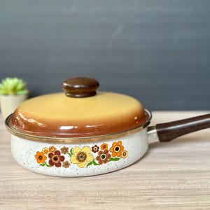 Puede incluir: Sartén vintage moteada blanca con mango y tapa marrones. La sartén presenta un diseño floral en tonos naranja, amarillo y marrón. La tapa tiene un degradado de amarillo a marrón. Un artículo de cocina clásico.