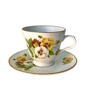 May include: A vintage teacup and saucer set. The white porcelain cup and saucer are adorned with a floral pattern of pansies in yellow, brown, and white, with a gold trim. The cup has a curved handle.