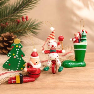 May include: A selection of wooden Christmas decorations. The ornaments include a green Christmas tree, a Santa Claus figure, a clown on a unicycle, and a Santa and tree in a green boot. Painted in bright colours.