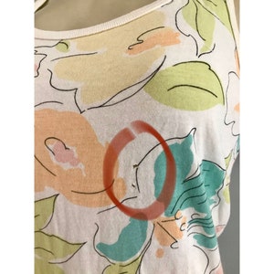 May include: A white tank top with a floral pattern in pastel colors. The design features large, stylized flowers in shades of peach, yellow, green, and teal, outlined in black. The neckline and straps are trimmed with a cream-colored fabric.