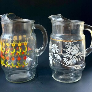 May include: Two glass pitchers with floral designs. One pitcher has a yellow, orange, and red floral design. The other pitcher has a white floral design with a gold band around the top.