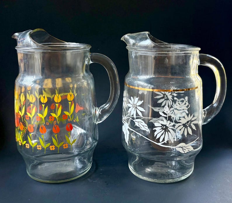 May include: Two glass pitchers with floral designs. One pitcher has a yellow, orange, and red floral design. The other pitcher has a white floral design with a gold band around the top.